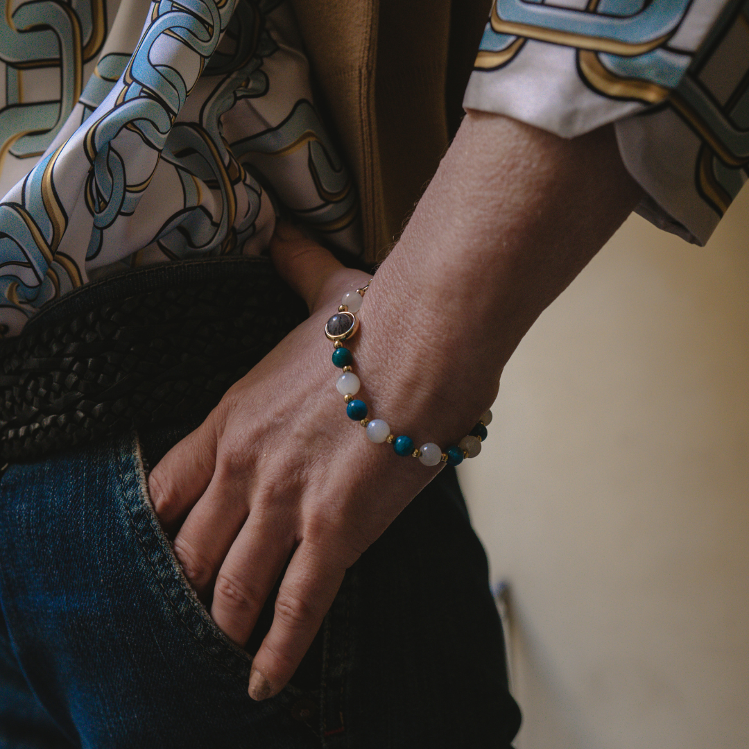 une Femme la mains dans sa poche mettant en avant son bracelet  Détoxifiant  composé de pierre naturelle d'Apatite bleue et Pierre de Lune , alternant avec de petites billes en or , avec une seule pierre de quartz rutile en son centre, ainsi qu'un noeud en or