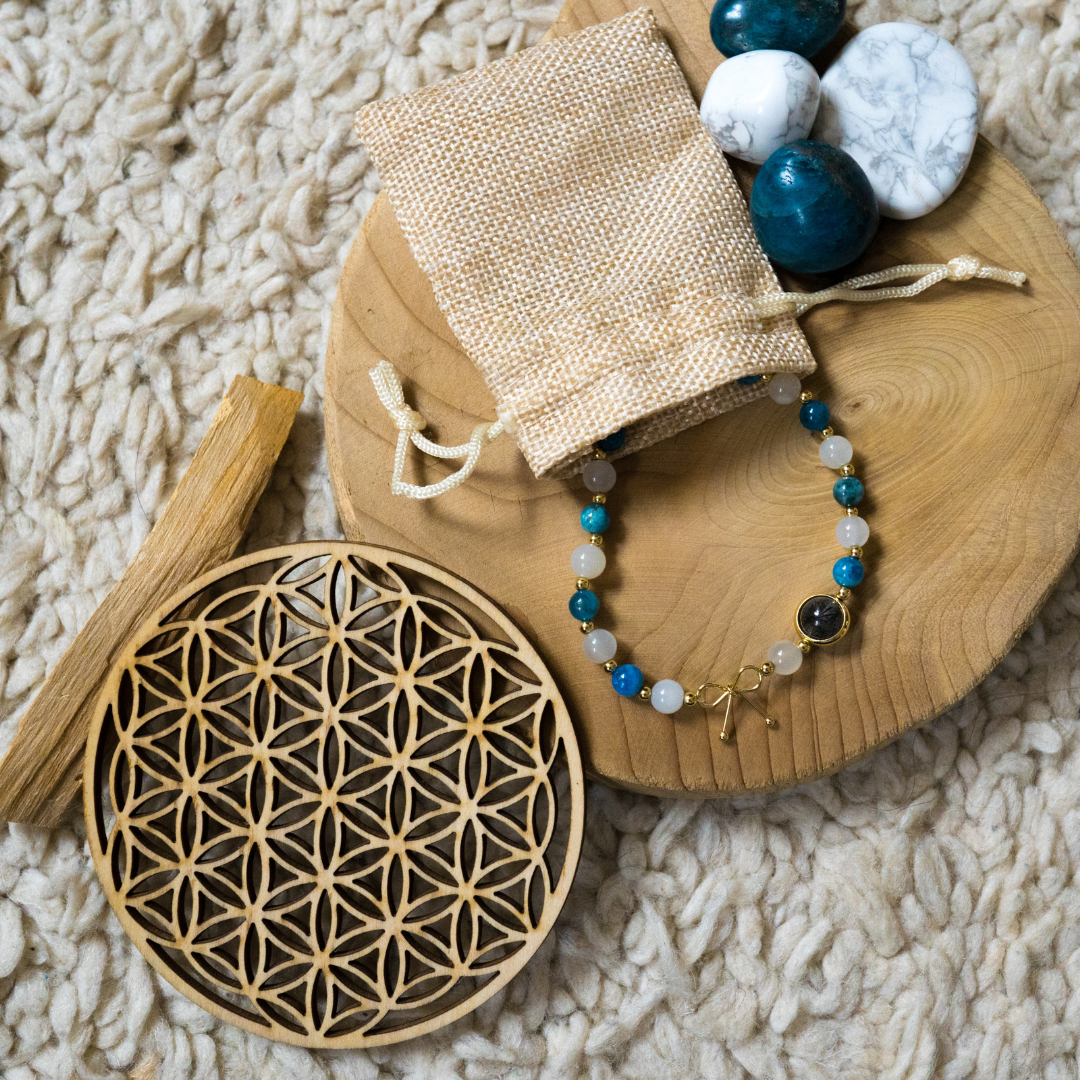 Bracelet  Détoxifiant composé de pierre naturelle d'Apatite bleue et Pierre de Lune , alternant avec de petites billes en or avec une seule pierre de quartz rutile en son centre, et son un noeud en or, posé sur un présentoir en bois quelques pierres d'apatite bleue , une fleur de vie ainsi que bâton à brûler de Palo santo